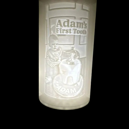 Decorative lamp with engraved design of a child and 'Adam's First Tooth' text, on a dark surface.